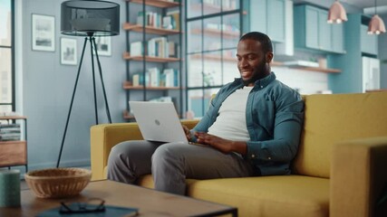 Handsome Black African American Man Working on Laptop Computer while Sitting on a Sofa in Cozy Living Room. Freelancer Working From Home. Browsing Internet, Using Social Networks, Having Fun in Flat.