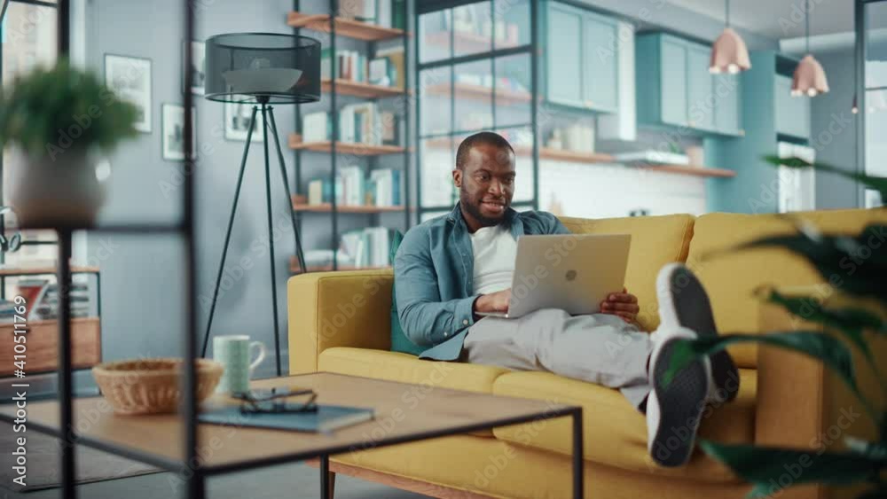 Aufkleber Handsome Black African American Man Working on Laptop Computer while Lying on a Sofa in Cozy Living Room. Freelancer Working From Home. Browsing Internet, Using Social Networks, Having Fun in Flat.