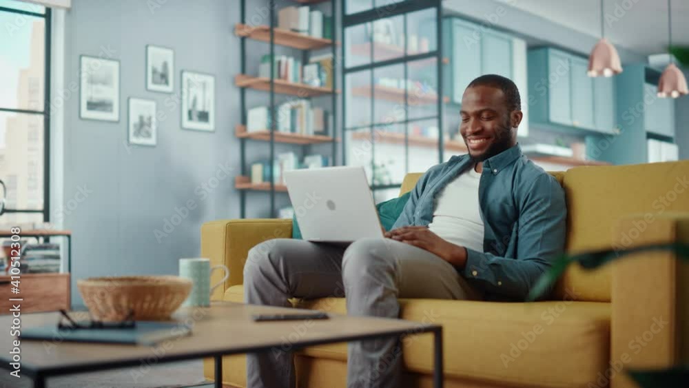 Poster happy black african american man working on laptop computer while sitting on a sofa in cozy living r