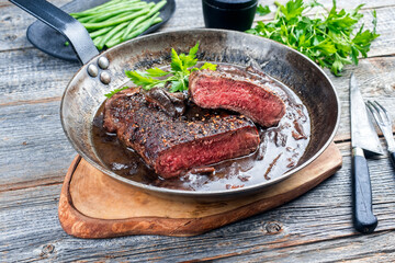 Traditionelle dry aged gebratenes Lendensteak vom Pferd mit Pilzen und Zwiebel in Rotwein Jus serviert als close-up in einer rustikalen Bratpfanne auf einem Holz Board