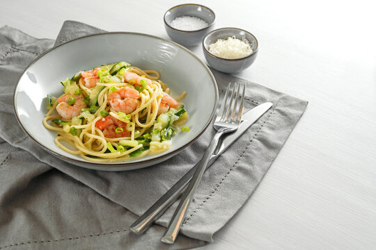 Spaghetti with zucchini courgettes, spring onions and prawns in a grey plate on a white table with a napkin and cutlery, healthy cooking concept, copy space