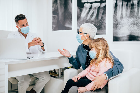Senior woman and her granddaughter sitting and talking with dentist at dentist's office. They are all wearing protective face masks due to coronavirus pandemic.