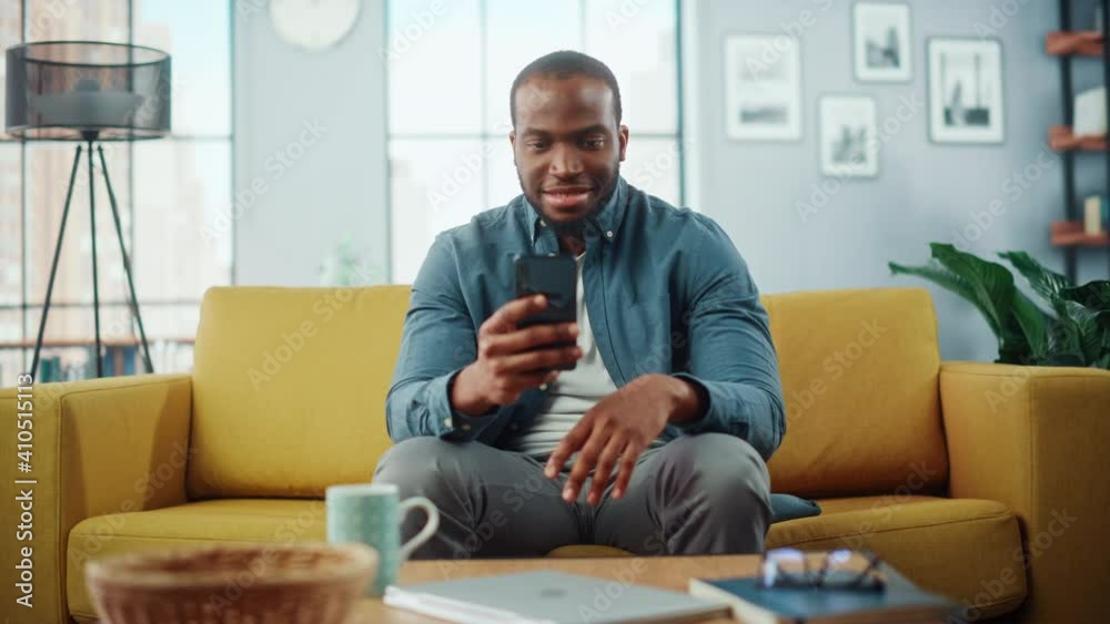 Aufkleber excited black african american man using smartphone while resting on a sofa in living room. happy ma