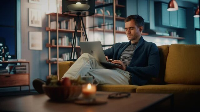 Handsome Caucasian Man Working on Laptop Computer while Sitting on a Sofa in Stylish Cozy Living Room. Freelancer Working From Home. Browsing Internet, Using Social Networks, Having Fun in Flat.