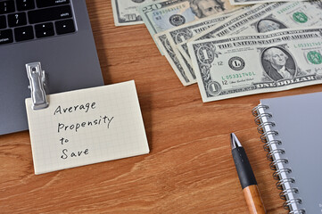 On the desk there are bills, a laptop, and memos with the word average propensity to save written on it.
