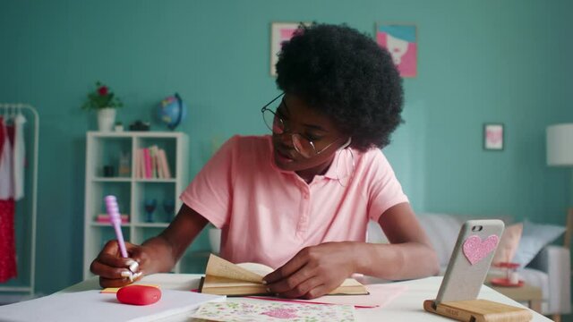 Young beautiful Afro-American woman, student, is studying at home, sitting at desk and reading interesting book, doing bookmarks to remember important details, foreground, Slow motion.