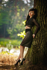 sexy brunette girl in a lace blouse and leather skirt with high heels posing near a tree in the...