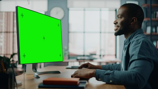 Handsome Black African American Specialist Working on Desktop Computer with Green Screen Mock Up Display at Home Living Room. Freelance Man Chatting to Clients Over the Internet on Social Networks.