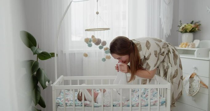 Cute adorable newborn baby girl lies on a back in the crib. A young beautiful mother plays with her little daughter, touches her feet. Concept of childhood, new life, parenthood.