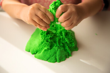 Close up of children's hands playing with green kinetic sand on white table; selective focus
