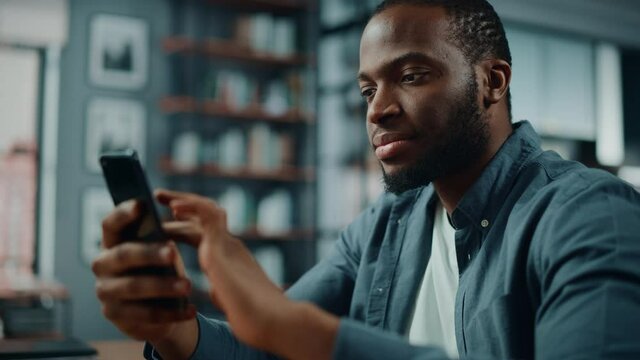 Excited Black African American Man Using Smartphone while Sitting at Table in Living Room. Happy Man Smiling at Home and Chatting to Colleagues and Clients Over the Internet. Using Social Networks.