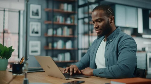 Handsome Black African American Man Working on Laptop Computer while Sitting Behind Desk in Cozy Living Room. Freelancer Working From Home. Browsing Internet, Using Social Network, Having Fun in Flat.