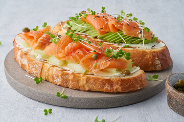 Two open sandwich, toast with salmon, cream cheese, avocado, cucumber slices on white concrete table. Morning healthy breakfast with fish and cup of drink, side view, close up