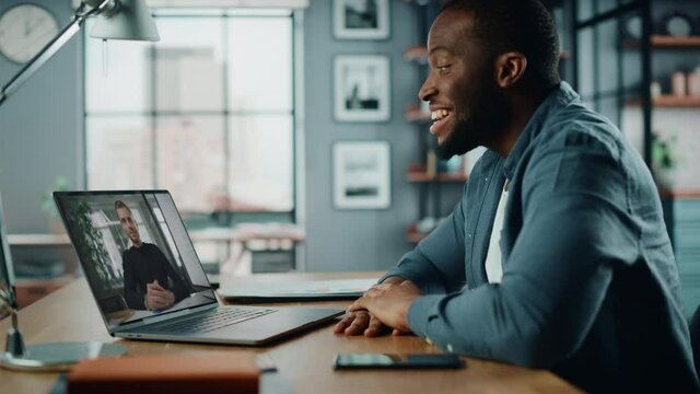 Handsome African American Man Having a Video Call on Laptop Computer while Sitting Behind Desk in Living Room. Freelancer Working From Home and Talking to Colleagues and Clients Over the Internet.