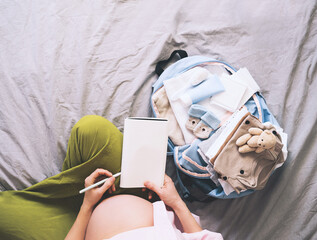 Pregnant woman packing hospital bag with checklist. Expectant mother with travel suitcase of baby clothes preparing for newborn birth, making notes in diary.