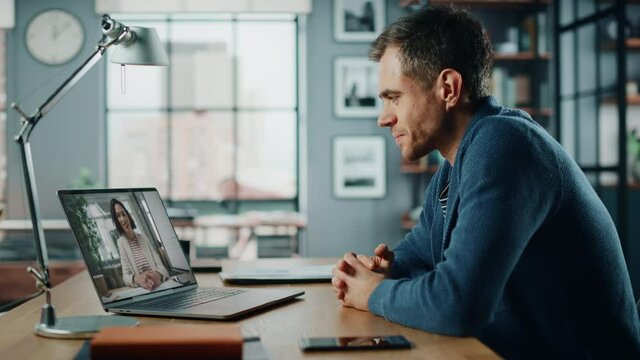 Handsome Caucasian Man Having a Video Call on Laptop Computer while Sitting Behind Desk in Living Room. Freelancer Working From Home and Talking to Colleagues and Clients Over the Internet.