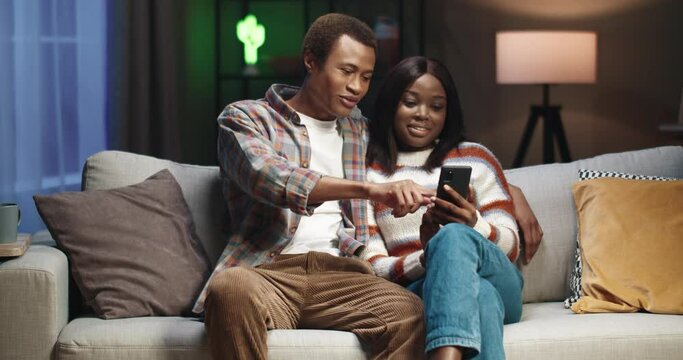 Young african american couple sitting at home on couch and flipping through the news feed on social networks using smartphone.