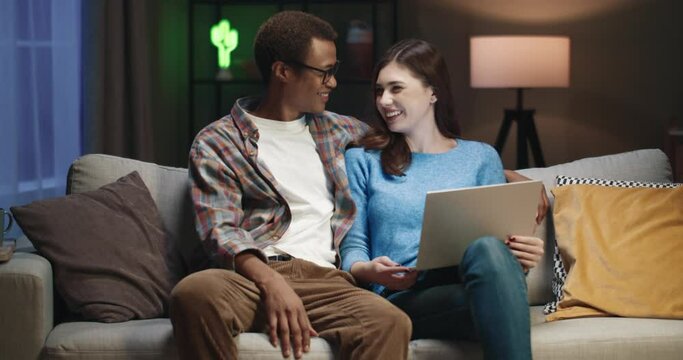 Young happy multinational couple sitting at home in evening on couch and spending free time watching interesting news on laptop.