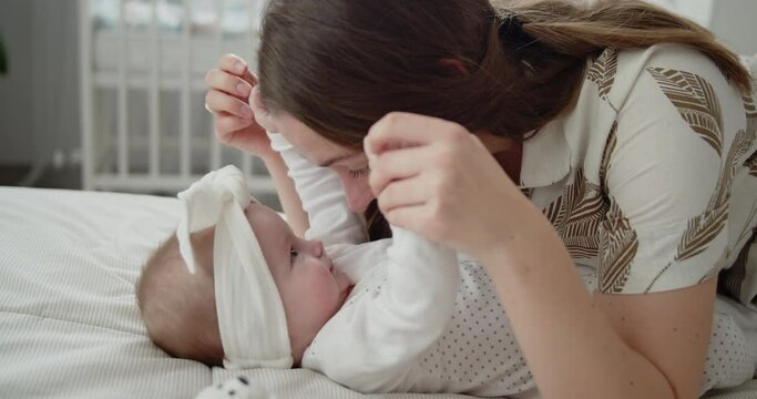 Young beautiful mother plays with her cute newborn baby that is lying on a back on a bed. Woman hold child's hands and kisses her. Concept of childhood, new life, parenthood.