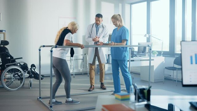 Hospital Physical Therapy: Strong Senior Female Disabled Patient with Injury Making First Steps, Walks Holding for Parallel Bars. Physiotherapist, Rehabilitation Doctor Applaud, Encourage Old Woman