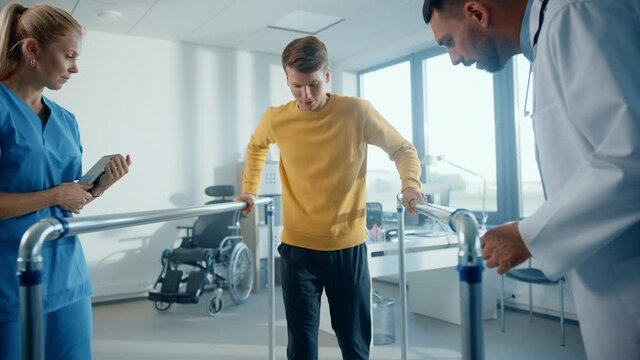 Hospital Physical Therapy: Determined Male Patient with Injury Successfully Walks Holding Parallel Bars. Physiotherapist, Rehabilitation Doctor Encourage, Applaud Disabled Person. Slow Motion