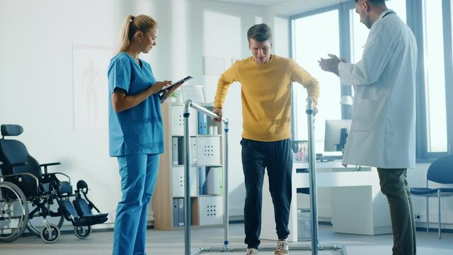 Hospital Physical Therapy: Strong Male Patient with Injury Successfully Walks Holding Parallel Bars. Physiotherapist, Rehabilitation Doctor, Help, Assist, Encourage Disabled Person. Slow Motion