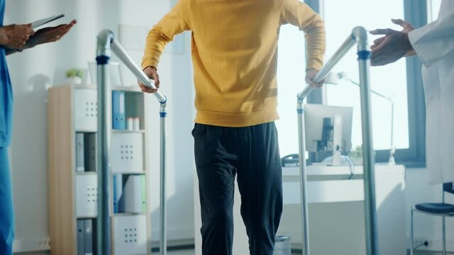 Hospital Physical Therapy: Portrait of Male Patient with Injury Successfully Walks Holding Parallel Bars. Physiotherapist, Rehabilitation Doctor, Help, Assist, Disabled Person. Elevating Slow Motion