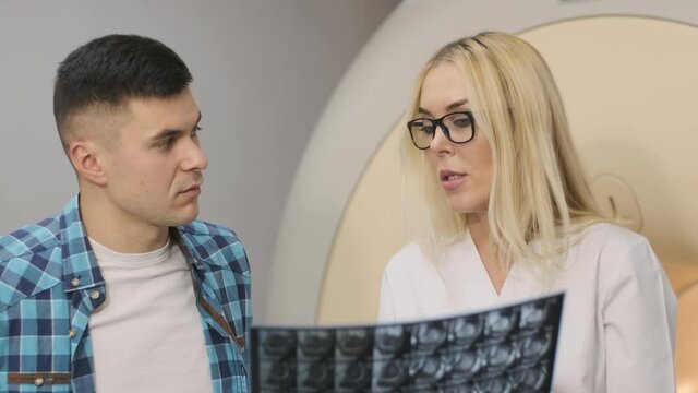 Woman doctor radiologist explains the results of MRI scanning for young male patient, showing the snapshot with images, observing and analyzing mri scan in modern clinic beside modern closed-type MRI