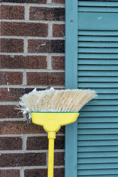 Broom Covered In Snow Leaning Against Brick Home With Blue Shutters; Cleanup After A Snow Storm