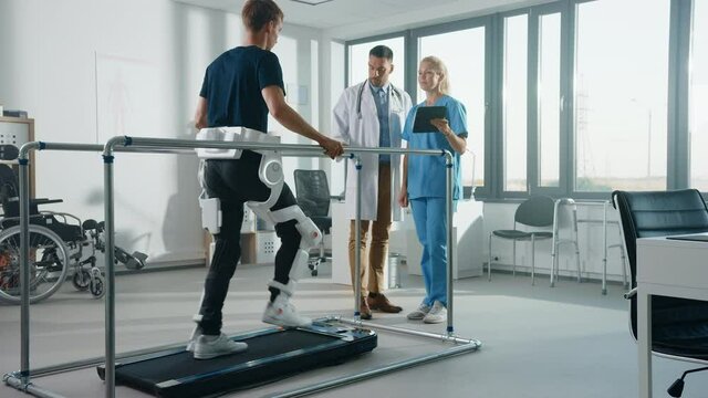 Modern Hospital Physical Therapy: Patient with Injury Walks on Treadmill Wearing Advanced Robotic Exoskeleton. Physiotherapy Rehabilitation Scientists, Engineers, Doctors use Tablet Computer to Help