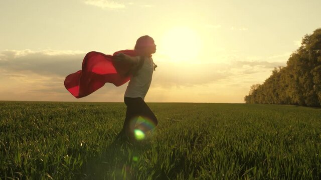 Child plays and dreams. Happy superhero girl, runs on green field in red cloak, cloak flutters in wind. Teenager dreams of becoming superhero. Young girl in red cloak, dream expression. Slow motion.