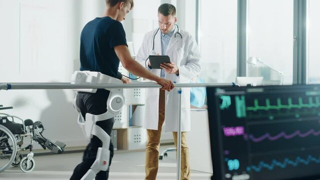 Modern Hospital Physical Therapy: Doctor Uses Tablet Computer, Disabled Patient Walks on Treadmill Wearing Advanced Robotic Exoskeleton. Computer Monitor Shows Physiotherapy Rehabilitation Activity