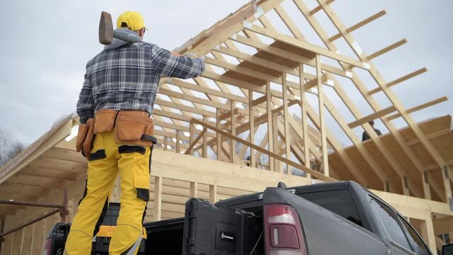 Construction Worker in His 40s with Large Hammer on His Shoulder. Wooden House Frame Structure in Background. Construction Zone Job.