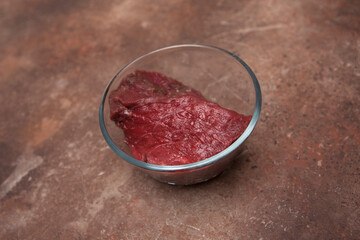 A piece of fresh raw beef in a transparent glass bowl on a granite table. Step by step recipe.