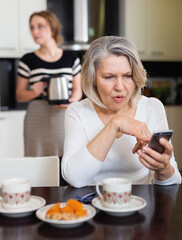 Upset mature mother using smartphone in kitchen interior while adult daughter making tea