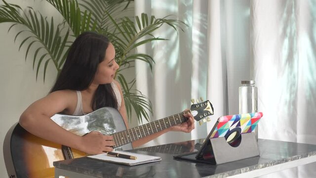 Pretty Indian girl watching online guitar lesson and practising music. Young Asian woman playing guitar at home. Aspiring musician learning to play musical instrument using laptop notepad.