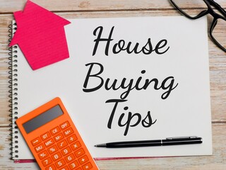 Property concept. Top view text House Buying Tips written on notebook with pen,calculator,magnifying glass,cupboard house,compass and eye glasses on wooden table.