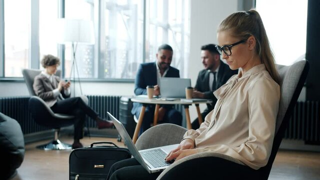 Attractive young businesswoman is working with laptop in hotel lobby while people are talking and walking with luggage in background. Business and travelling concept.