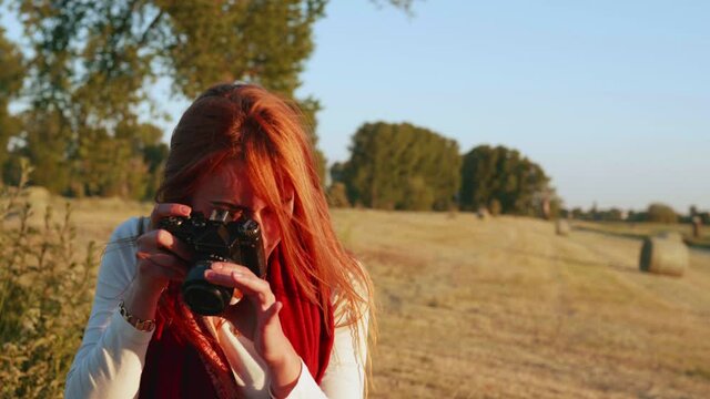 [4k] slow motion portrait of beautiful long hair ginger woman taking a picture on a meadow with hay barns in background and hair blowing in the wind