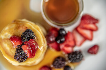 Tasty, yummy breakfast of pancakes stacked with  strawberries, raspberries and black berries placed on a white plate surrounded by maple syrup, honey. Taken in professional studio setting.  
