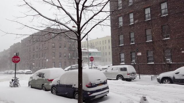 A slow-motion pan to the right shows heavy snowfall and piles of snow covering cars in a New York area neighborhood.