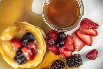 Tasty, yummy breakfast of pancakes stacked with  strawberries, raspberries and black berries placed on a white plate surrounded by maple syrup, honey. Taken in professional studio setting.  