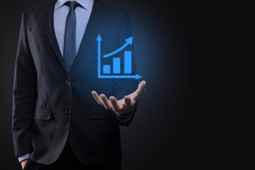 Businessman holding tablet and showing a growing virtual hologram of statistics, graph and chart with arrow up on dark background. Stock market. Business growth, planing and strategy concept.