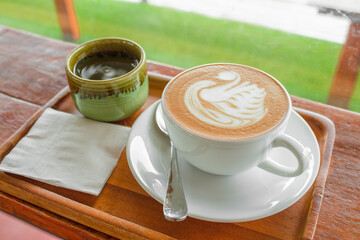 A cup of coffee with latte art in a swan shape on a wooden table, White hot coffee mug with green tea cup.