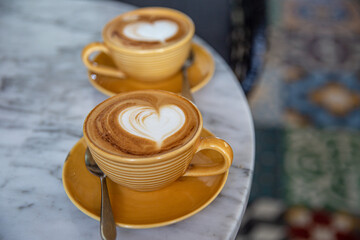 Two yellow cups of hot cappuccino on marble table background. Heart shape as art latte for love