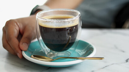 Asian woman hand with a cup of black coffee on blue plate with golden spoon, homemade americano good for healthy lifestyle on relaxing day, isolated in white granite table. Classy coffee shop concept.