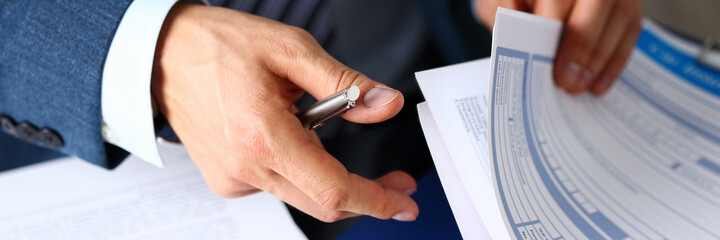 Male arm in suit offer insurance form clipped to pad and silver pen to sign closeup. Strike a...