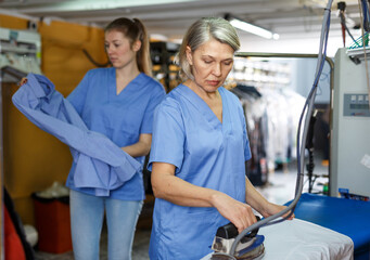 Two female cleaning services salon employees in process of ironing clean clothes