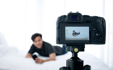 Asian young blogger man playing internet smartphone on his white bed after mockup focus position for record live video promote product by camera with tripod at bedroom