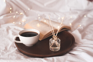 Cup of fresh black coffee with open paper book and liquid home perfume in glass bottle with sticks over glowing lights in bed close up. Cozy atmosphere. Good morning.